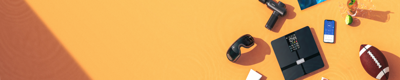 A collection of items on an orange surface, including a mobile phone, football, sunglasses, scale, miniature weight, goggles and some other exercising equipment.