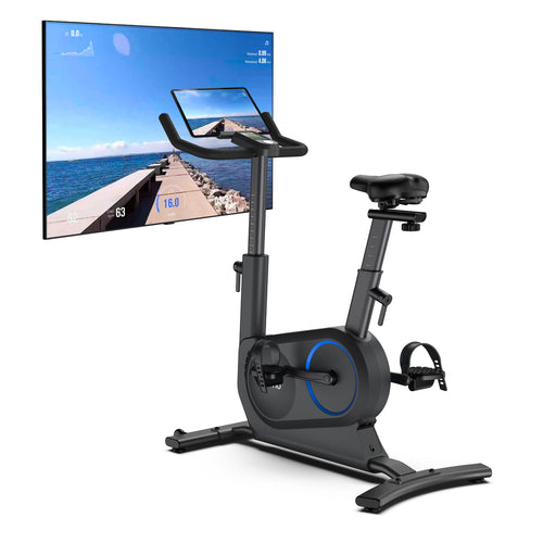 A high-tech AI Smart Bike S by Renpho UK featuring a tablet screen mounted on the handlebars is shown. The bike is positioned in front of a large wall-mounted flat-screen TV displaying a scenic road view with fitness data overlay. Perfect for indoor cycling, it has adjustable handlebars, seat, and pedals with straps. (A)